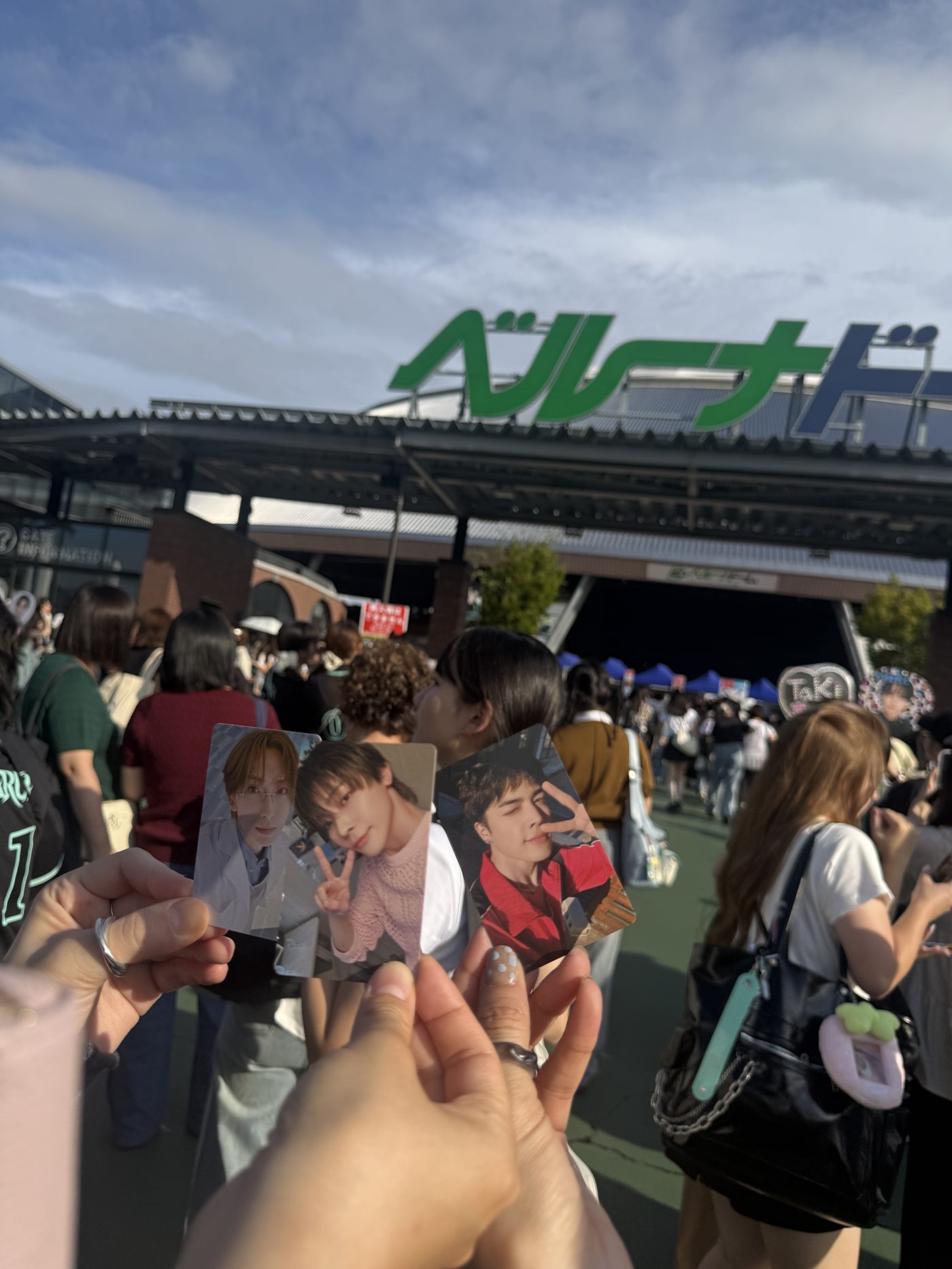 Fans holding up &TEAM member photocards under a bright blue sky in front of the green Belluna Dome sign on arrival