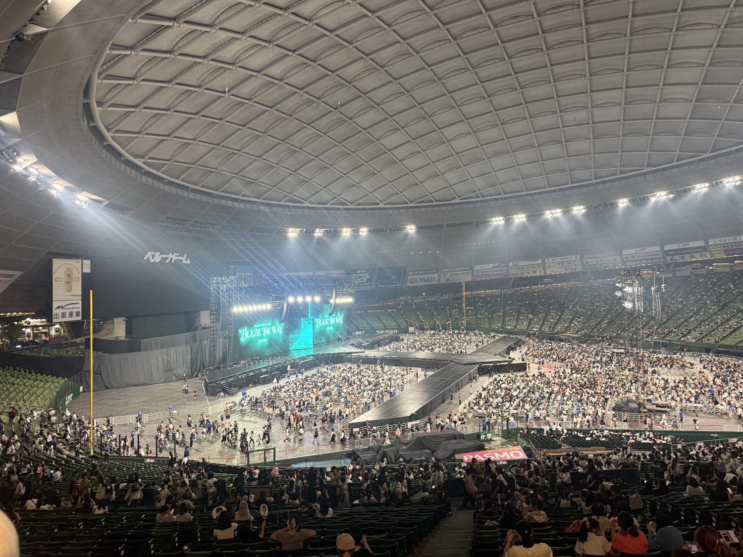 Belluna Dome at night during the concert, stage lit teal under spotlights with the BLAZE THE WAY screens and a huge crowd across the floor and stands