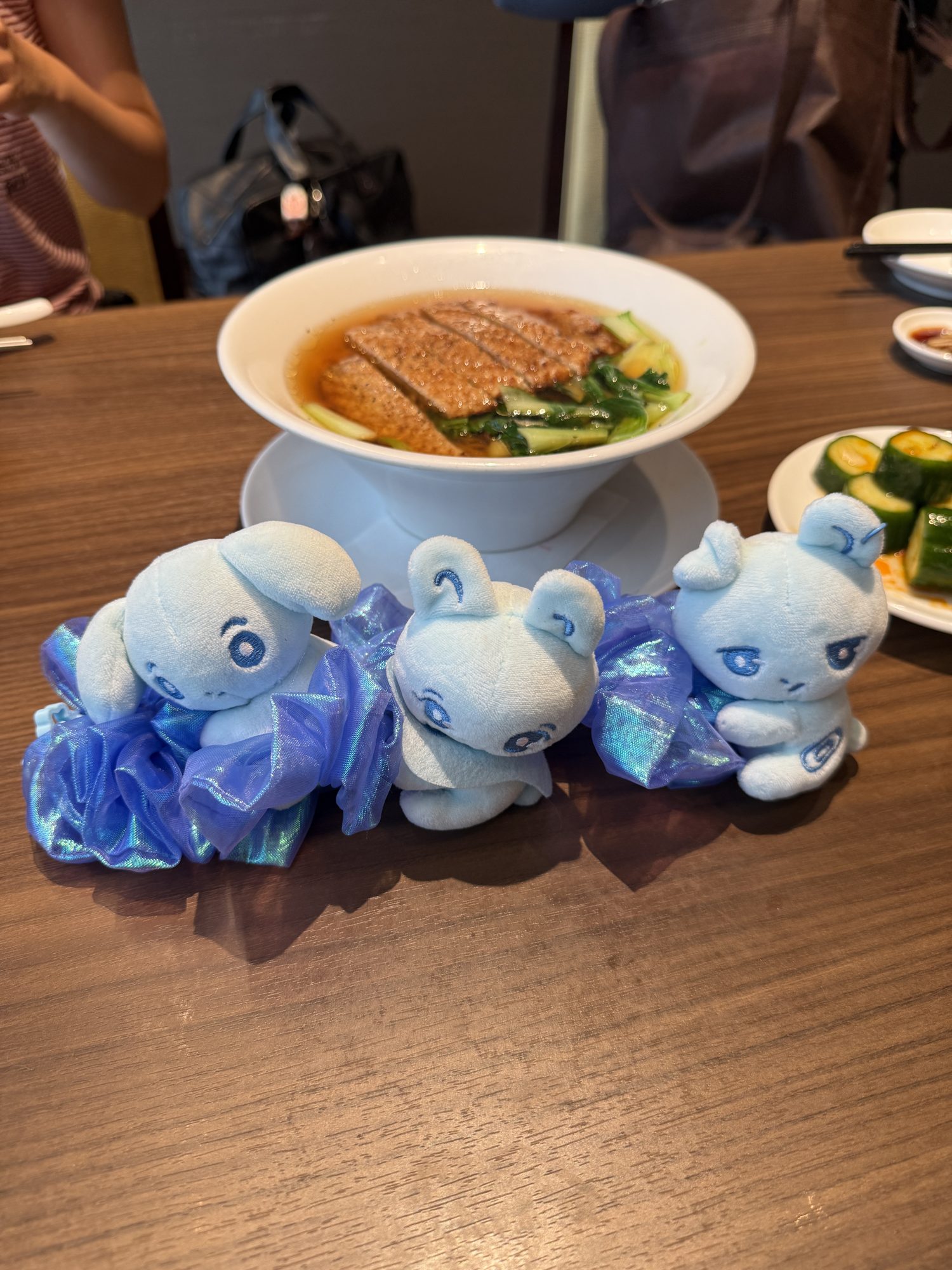 Three pale-blue &TEAM LUNE character plushies lined up in front of a bowl of sliced meat over greens at a restaurant table