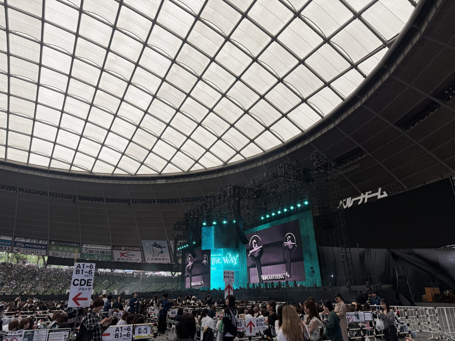 The white latticed roof of Belluna Dome curving over the lit concert stage under a flat grey sky through the open sides