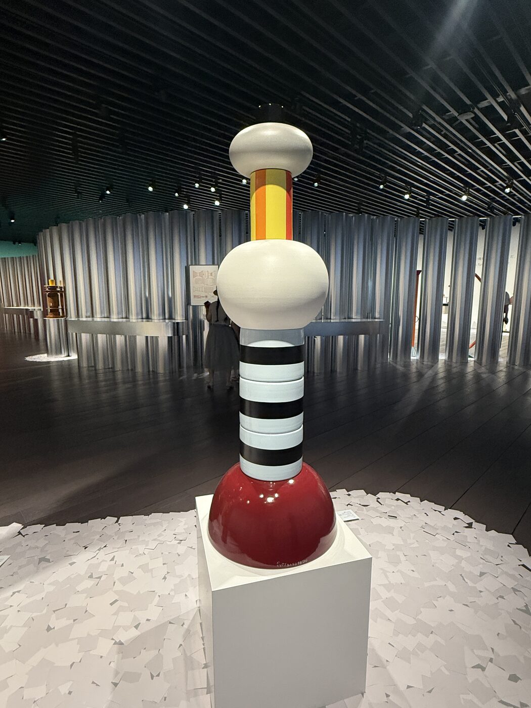 A tall totem-like Sottsass ceramic on a plinth in a room of vertical light columns