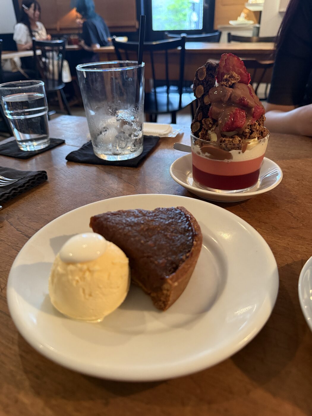 A tart with vanilla ice cream, and a berry parfait behind, on a wooden table