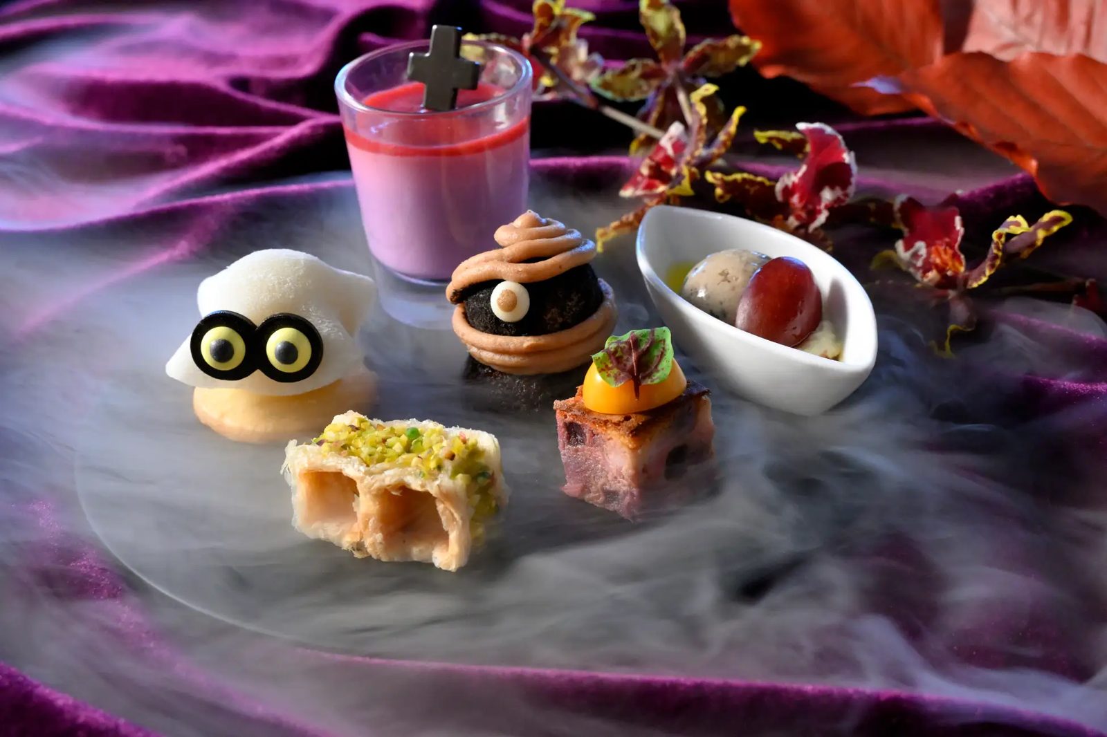 Close-up of Halloween petits fours — a ghost, a blood-red cross verrine, a chocolate choux and an autumn-leaf cube — on purple velvet with mist