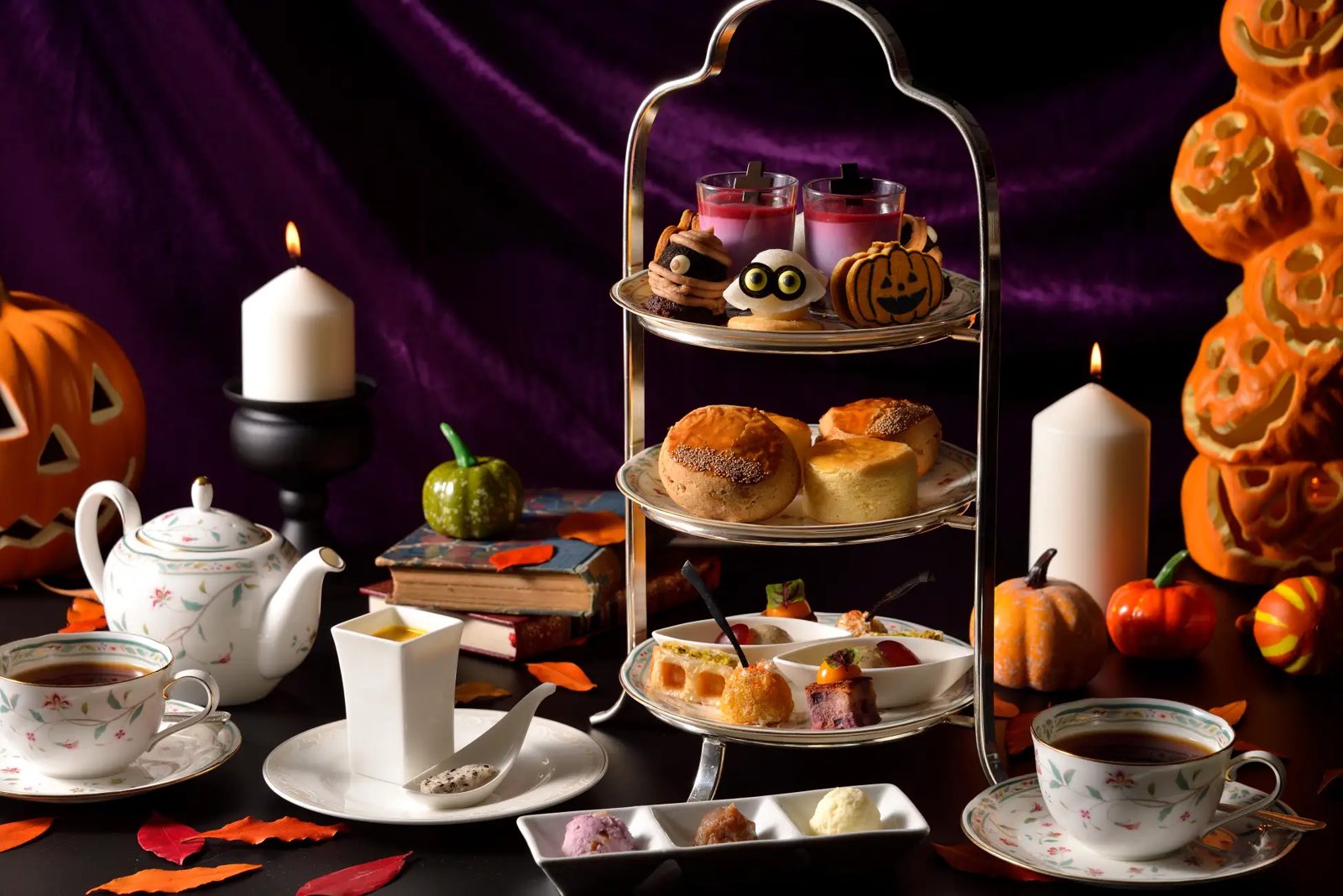 A full three-tier afternoon tea stand with floral china, teapot, candles and carved pumpkins against a purple drape