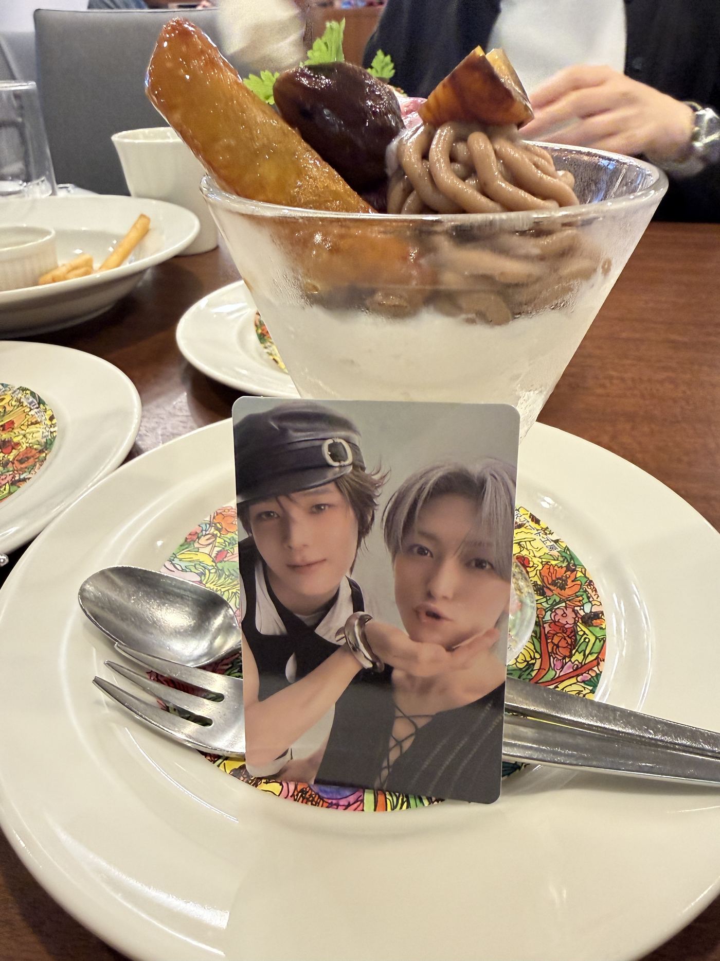 A wide glass parfait topped with a long candied sweet potato, a glazed chestnut and piped marron cream, with a K-pop photocard propped against the spoon on the plate in front