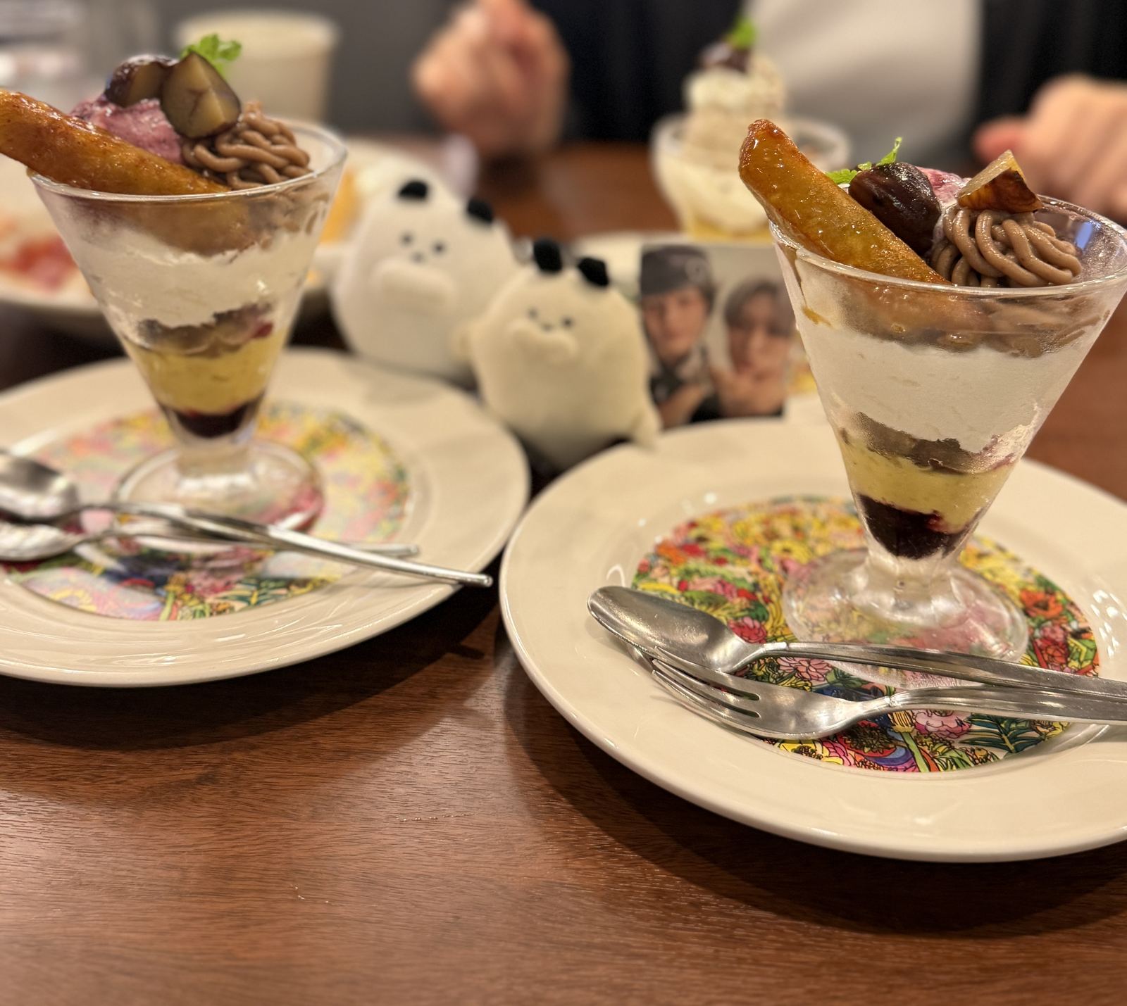 Two Royal Host autumn parfaits in stemmed glasses, topped with candied sweet potato, chestnut and piped marron cream over layers of cream, sweet potato and dark berry sauce, with two small white plush toys and a photocard on the table between them