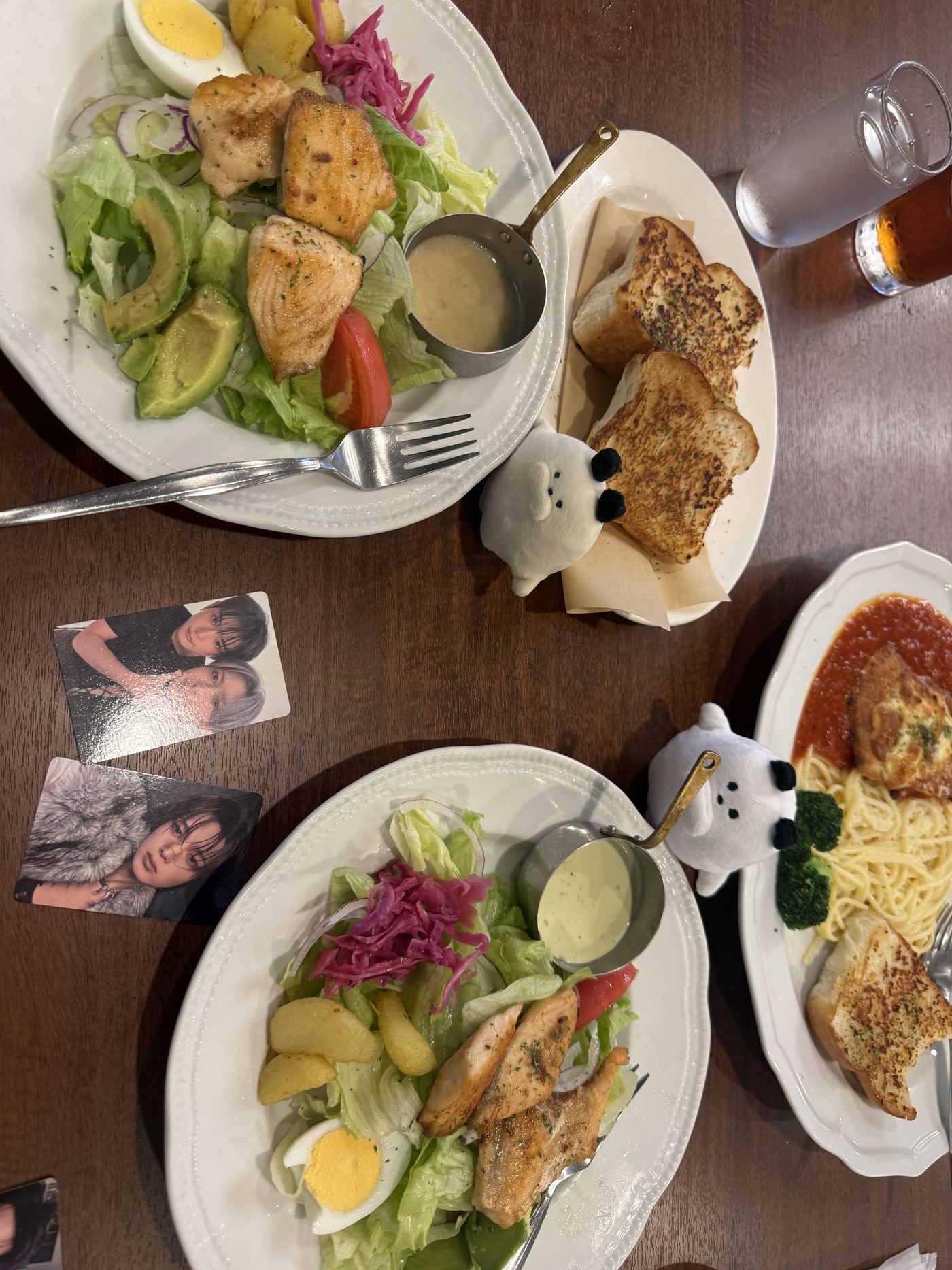 Overhead view of a Royal Host table with two salads topped with grilled chicken, avocado, egg and pickled red cabbage, toasted garlic bread, a tomato-sauce pasta dish, two small white plush toys and photocards