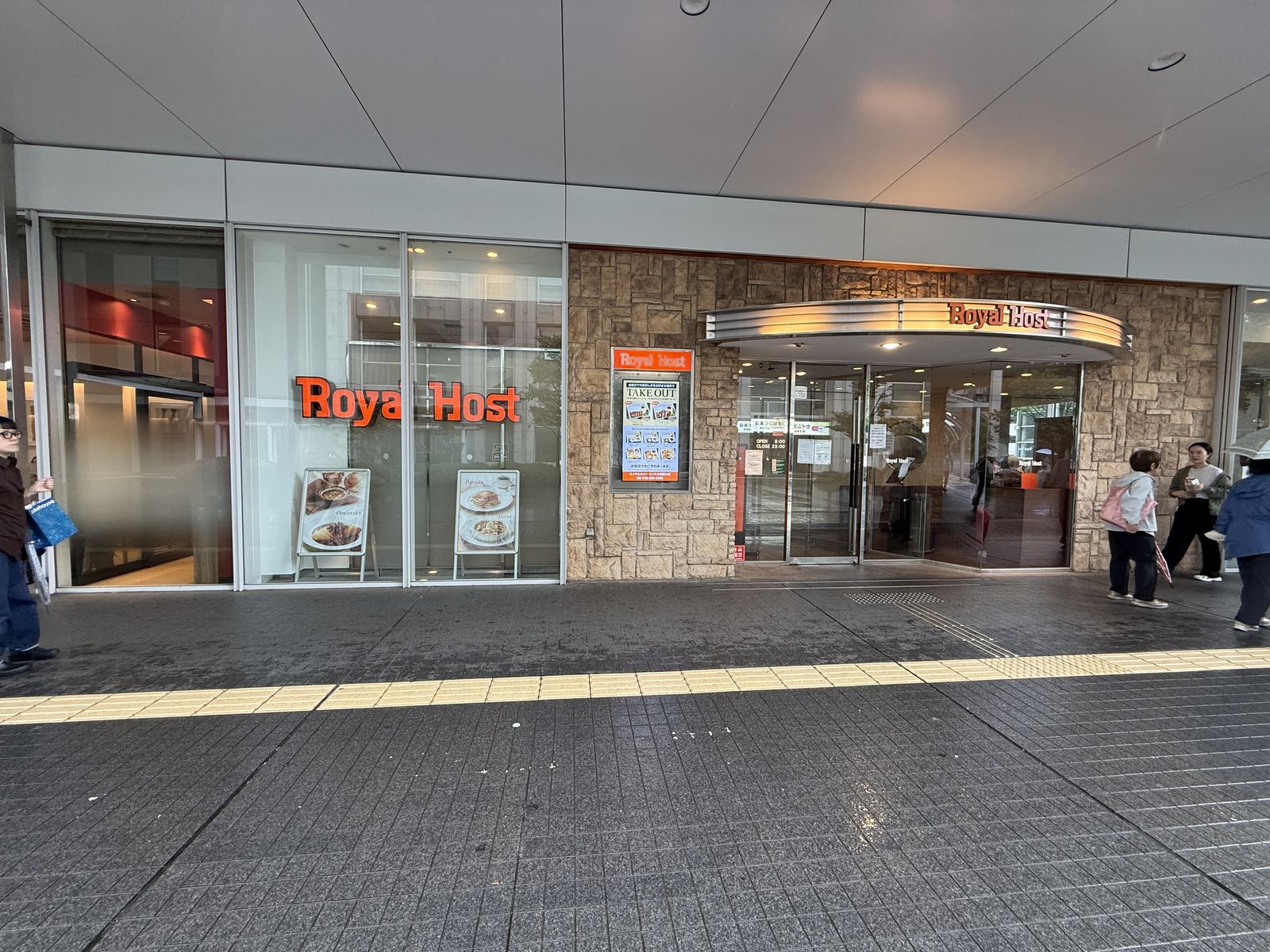 The street-level entrance of Royal Host Saitama-Shintoshin, with the red Royal Host lettering on the glass, a stone-clad wall with a digital menu board, and a covered walkway in front