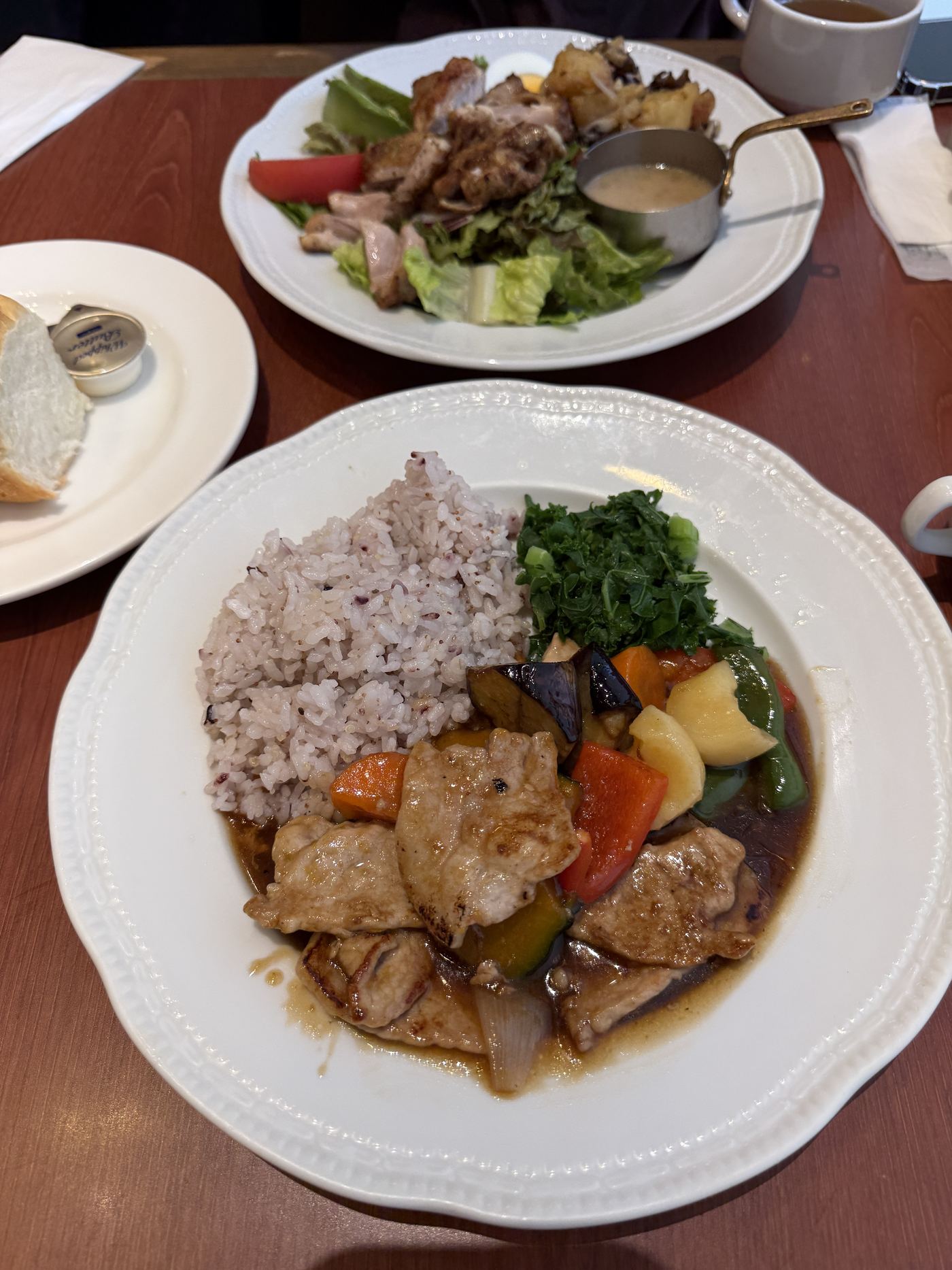 A Royal Host rice plate of multigrain rice, sautéed pork and seasonal vegetables in a brown sauce with greens, with a side salad and bread behind it
