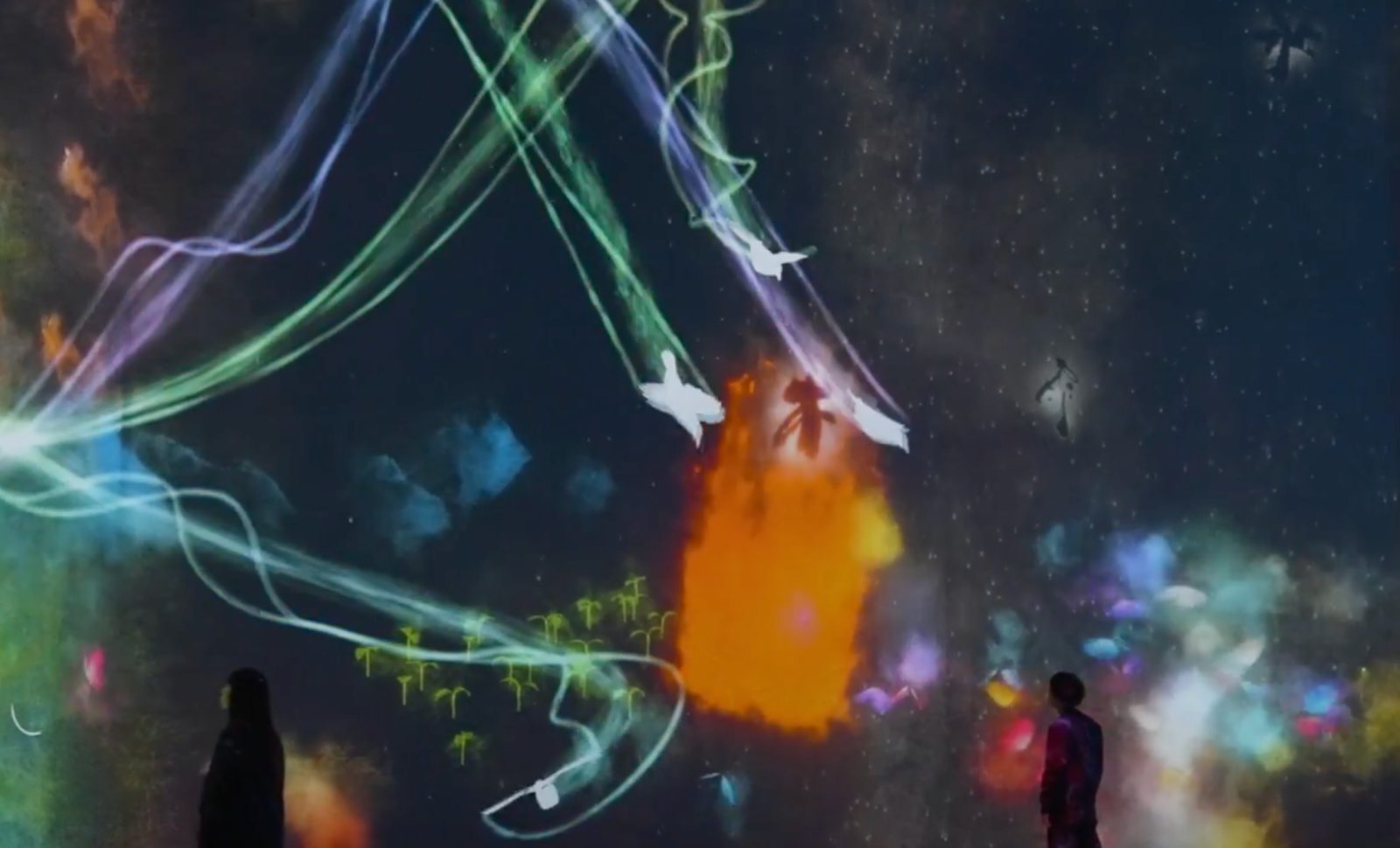 Streams of coloured light, white birds and drawn creatures spread across a dark wall as two visitors stand watching in silhouette at teamLab Borderless
